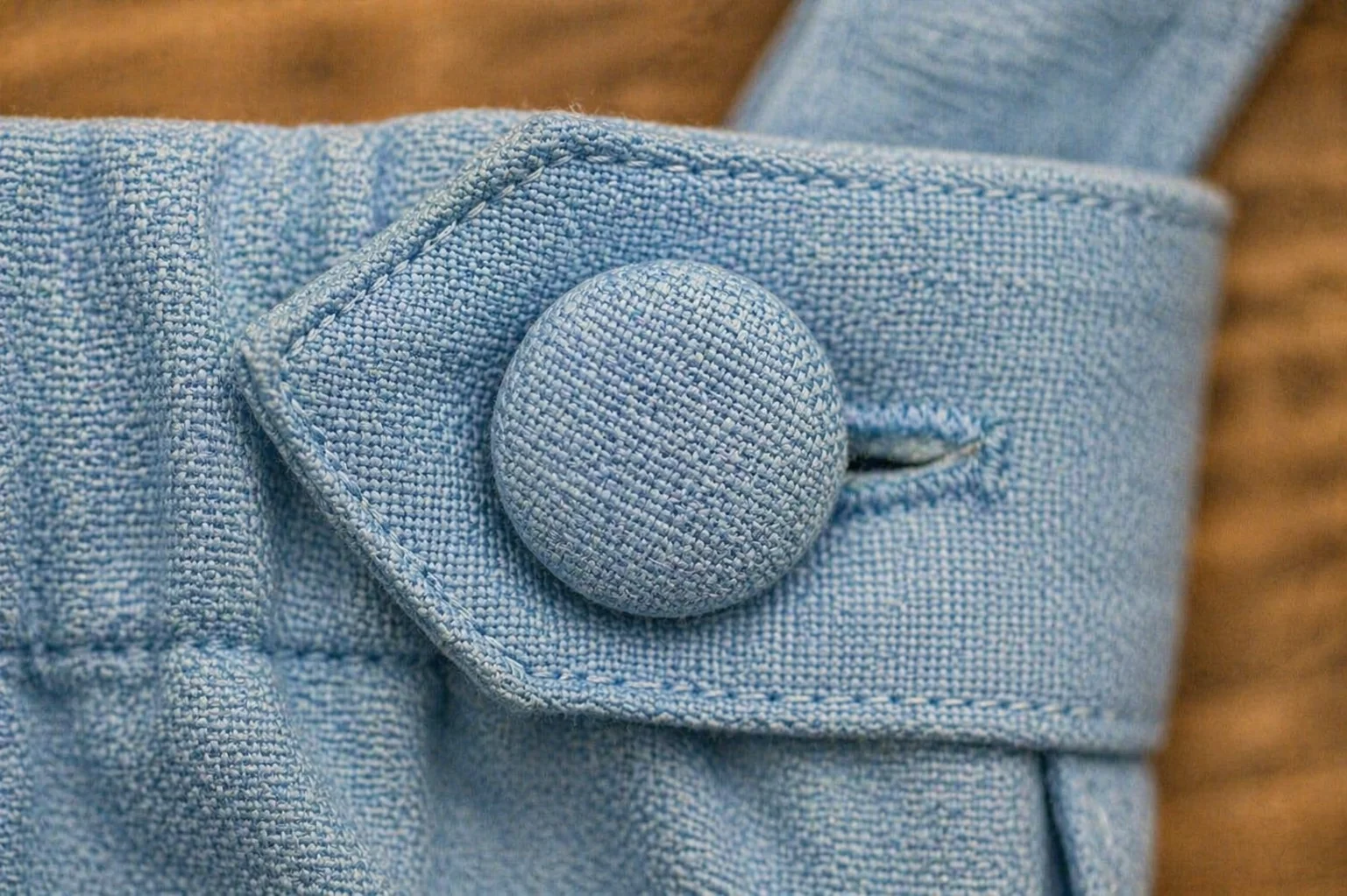 Macro extremo de botão encapado no cós lateral da Jardineira Versátil · botão coberto com o próprio tecido linho azul céu pastel · casa de botão funcional com costura limpa · zero plástico ou metal visível · alternativa premium ao botão de pressão