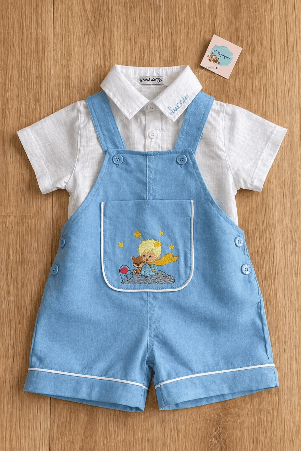 Jardineira Versátil flat-lay editorial — peça em azul céu linho com bordado Pequeno Príncipe no bolso, body camisa branca e tag @ateliedagiinfantil