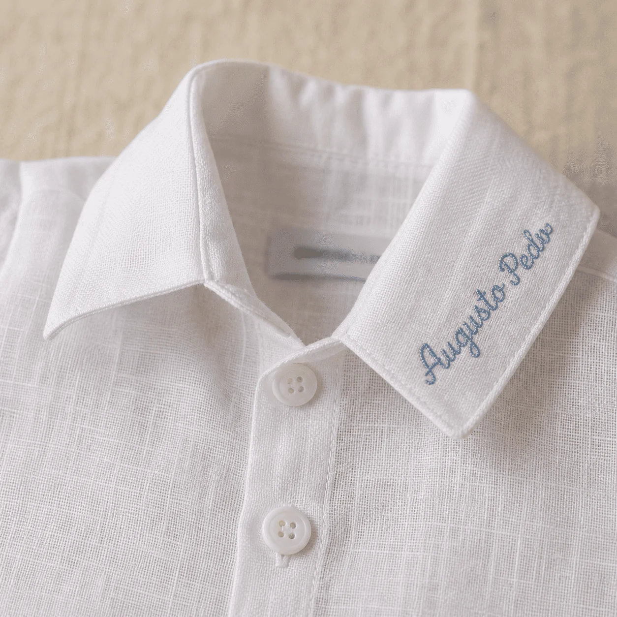 Macro do colarinho do body social branco com o nome 'Augusto Pedro' bordado em script azul-claro — gola estruturada por fora, leve por dentro.